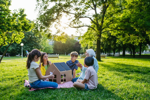 Children learning about renewable solar energy during sustainable education class outdoors, using cardboard model of house wit solar panel on roof.