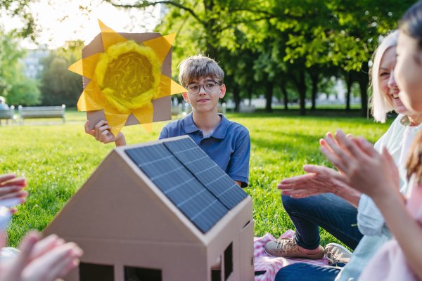 Children learning about renewable solar energy during sustainable education class outdoors, using cardboard model of house wit solar panel on roof.