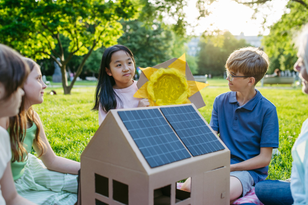 Children learning about renewable solar energy during sustainable education class outdoors, using cardboard model of house wit solar panel on roof.