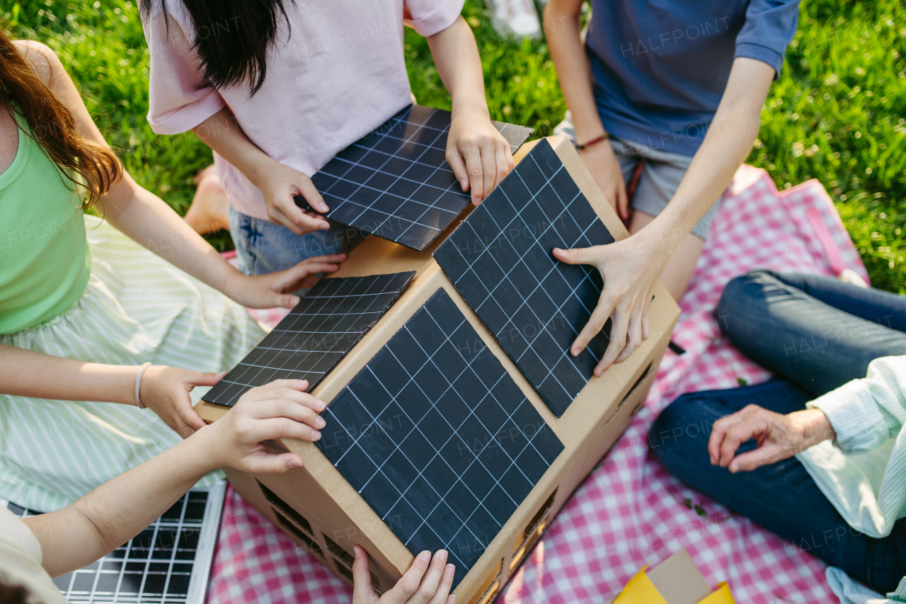 Children learning about renewable solar energy during sustainable education class outdoors, using cardboard model of house wit solar panel on roof.