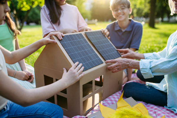 Children learning about renewable solar energy during sustainable education class outdoors, using cardboard model of house wit solar panel on roof.