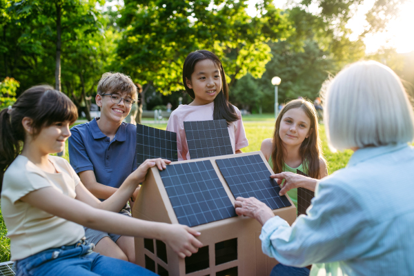 Children learning about renewable solar energy during sustainable education class outdoors, using cardboard model of house wit solar panel on roof.