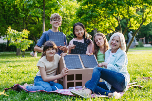 Children learning about renewable solar energy during sustainable education class outdoors, using cardboard model of house wit solar panel on roof.