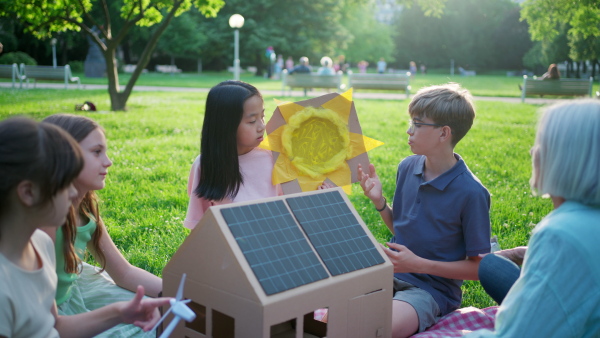 Children learning about renewable solar energy during sustainable education class outdoors, using cardboard model of house wit solar panel on roof.