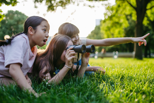 Young girl students learning about nature and wildlife in urban environment, using binoculars and observing animals, birds in public park.