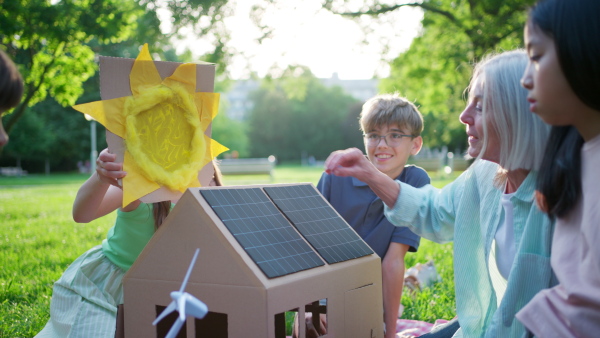 Children learning about renewable solar energy during sustainable education class outdoors, using cardboard model of house wit solar panel on roof.