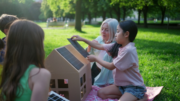 Children learning about renewable solar energy during sustainable education class outdoors, using cardboard model of house wit solar panel on roof.