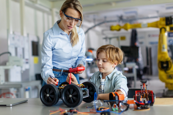 Female teacher helping young schoolboy to build robotic car in after-school robotics club. Children learning robotics in Elementary school. Science for kids. STEM.