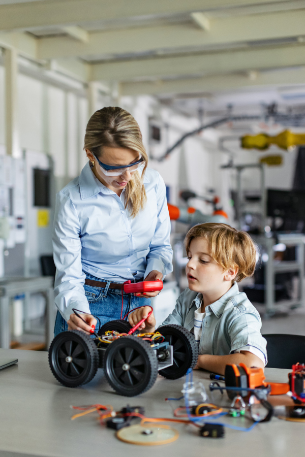 Female teacher helping young schoolboy to build robotic car in after-school robotics club. Children learning robotics in Elementary school. Science for kids. STEM.