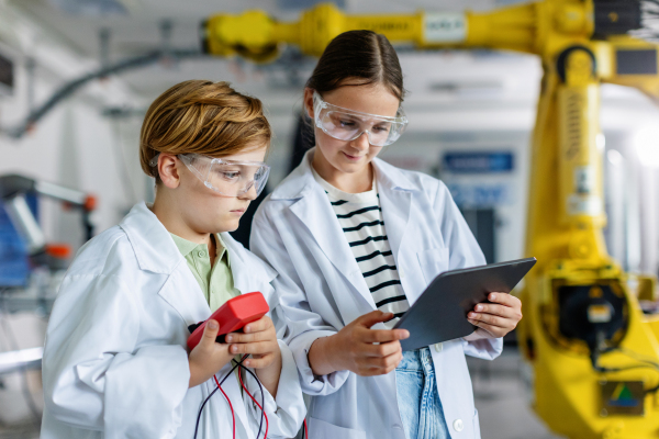 Two classmates standing in a robotic laboratory, wearing lab coats and safety eyeglasses. After-school robotics club. Children learning robotics in Elementary school.