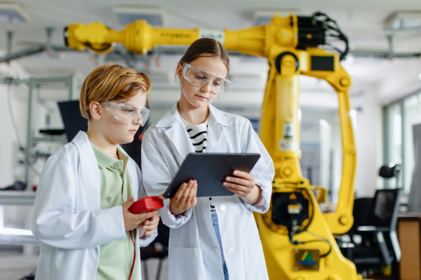 Two classmates standing in a robotic laboratory, wearing lab coats and safety eyeglasses. After-school robotics club. Children learning robotics in Elementary school.