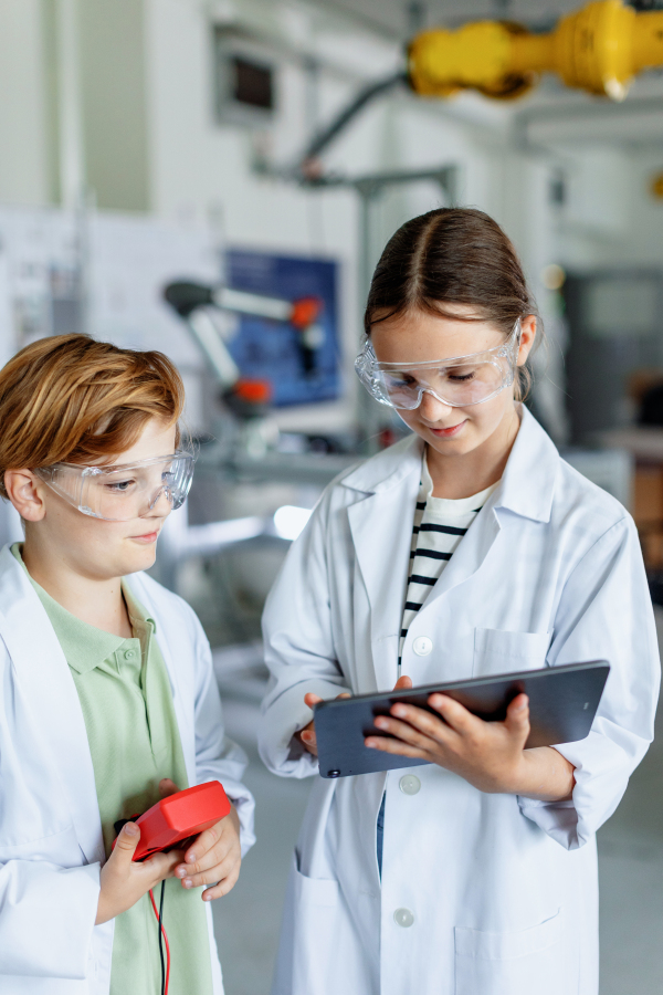 Two classmates standing in a robotic laboratory, wearing lab coats and safety eyeglasses. After-school robotics club. Children learning robotics in Elementary school.