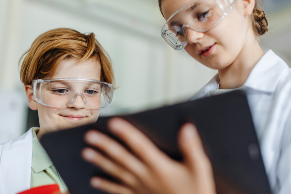 Two classmates standing in a robotic laboratory, wearing lab coats and safety eyeglasses. After-school robotics club. Children learning robotics in Elementary school.