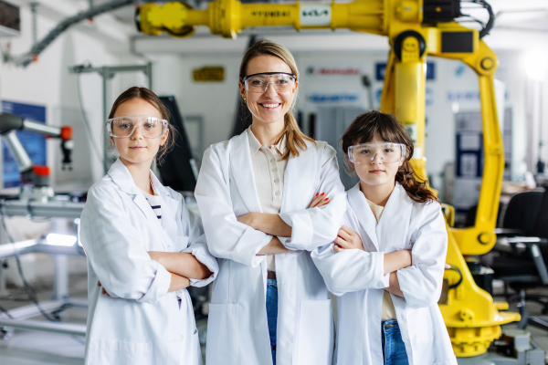 Female teacher with two schoolgirls in lab coats, standing in front robotic arm. After-school robotics club. Children learning robotics in Elementary school. Girls in science. STEM.