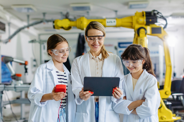 Female teacher with two schoolgirls in lab coats, standing in front robotic arm. After-school robotics club. Children learning robotics in Elementary school. Girls in science. STEM.