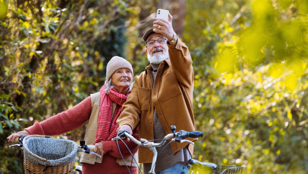 Lovely elderly couple on standing in the middle of autumn nature with their bikes, taking selfie with smartphone