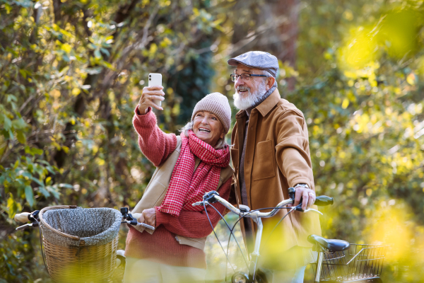 Lovely elderly couple on standing in the middle of autumn nature with their bikes, taking selfie with smartphone