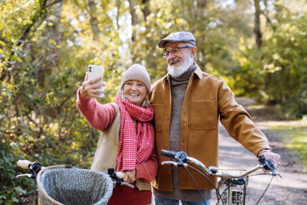 Lovely elderly couple on standing in the middle of autumn nature with their bikes, taking selfie with smartphone