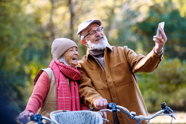 Lovely elderly couple on standing in the middle of autumn nature with their bikes, taking selfie with smartphone