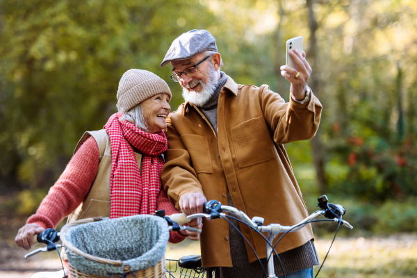 Lovely elderly couple on standing in the middle of autumn nature with their bikes, taking selfie with smartphone