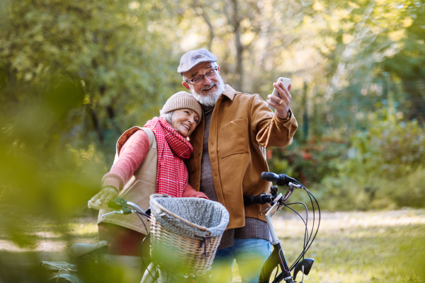 Lovely elderly couple on standing in the middle of autumn nature with their bikes, taking selfie with smartphone