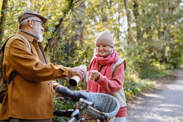 Senior couple with bikes standing in the middle of autumn nature and resting, enjoying cup of hot coffee or tea from thermos.