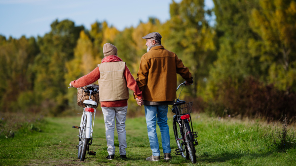 Rear view of elderly couple on walk in the forest, pushing their bikes side by side. Seniors in love on stroll through autumn nature, enjoying a peaceful moment together.