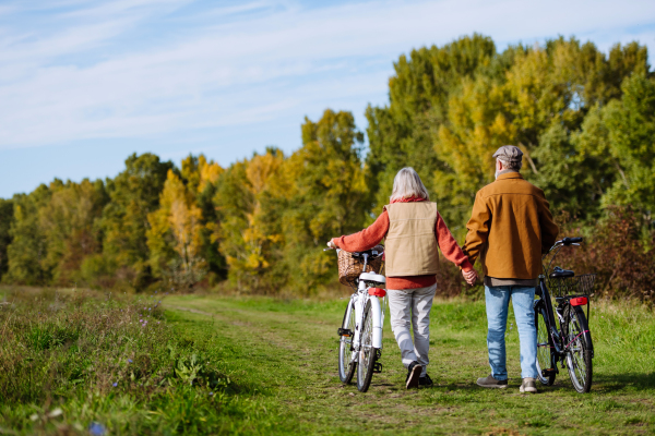 Rear view of elderly couple on walk in the forest, pushing their bikes side by side. Seniors in love on stroll through autumn nature, enjoying a peaceful moment together.