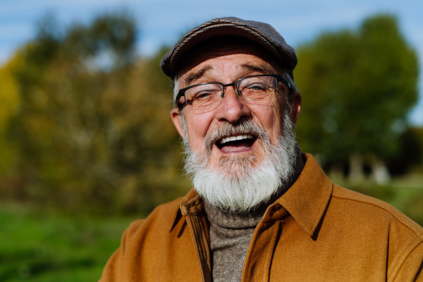 Portrait of friendly older man with gray beard. Senior standing outdoors in nature, looking at camera and smiling.