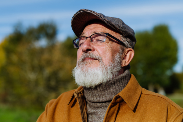 Portrait of handsome older man with a gray beard. Senior standing outdoors in nature, enjoying the sun's rays on his face