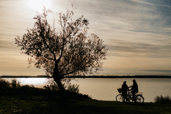 Lovely elderly couple on standing by lake with their bikes, looking at setting sun, enjoying peaceful moment together.