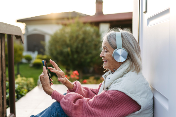 Elderly woman with gray hair listening music outdoor. Female senior wearing headphones and playing music from phone.