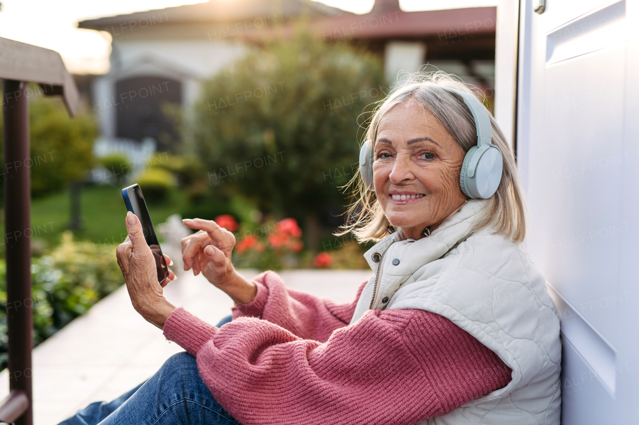 Elderly woman with a gray hair listening music outdoor. Female senior wearing headphones and playing music from phone.