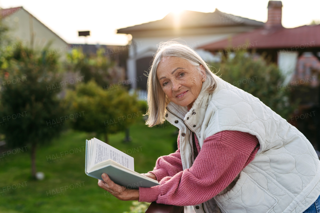 Elderly woman standing on balcony, holding book and reading. Side view. Female senior enjoying calm moment with good book.