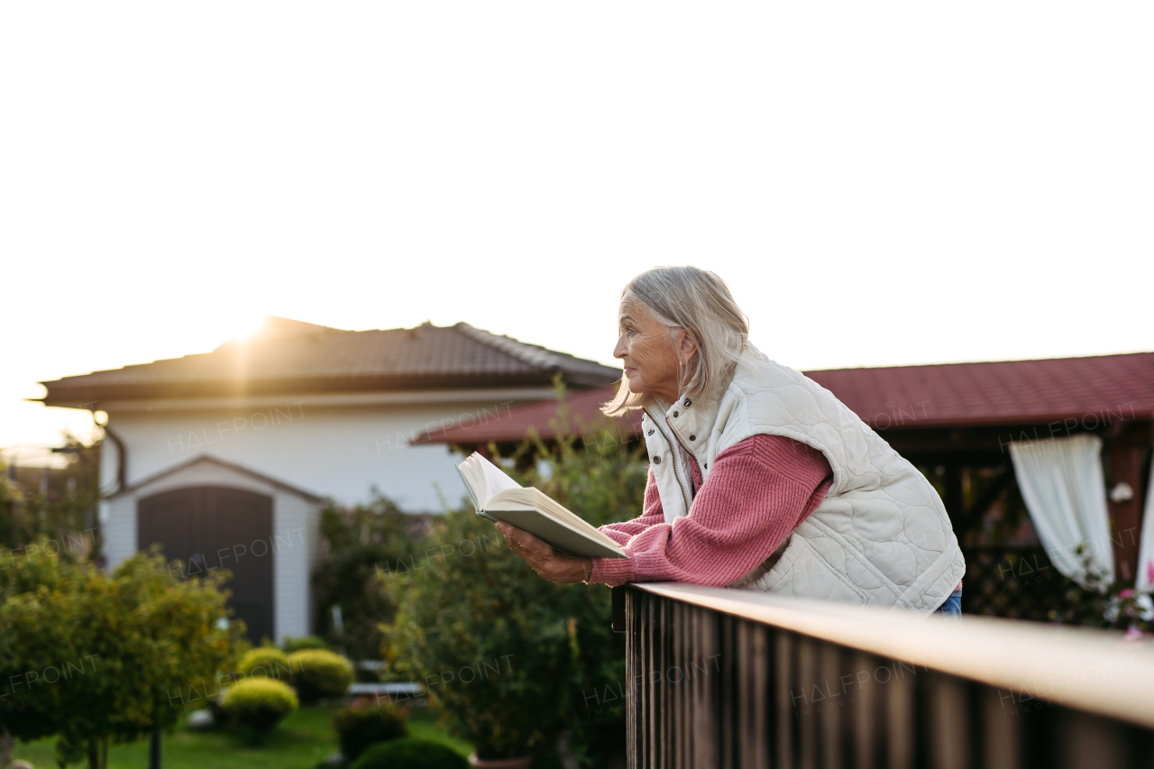 Elderly woman standing on balcony, holding book and reading. Side view. Female senior enjoying calm moment with good book.