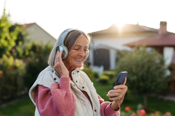 Elderly woman with a gray hair listening music outdoor. Female senior wearing headphones and playing music from phone.