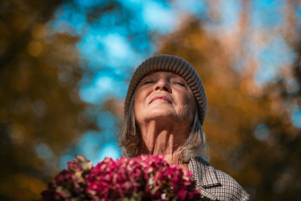 Portrait of beautiful senior woman standing outdoors with her eyes closed. Autumn nature behind her.