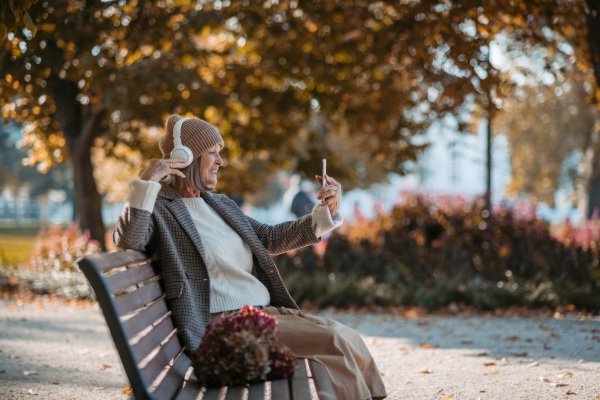 Portrait of a beautiful senior woman with headphones in autumn park, listening music and holding phone.