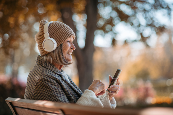 Portrait of a beautiful senior woman with headphones in autumn park, listening music and holding phone.