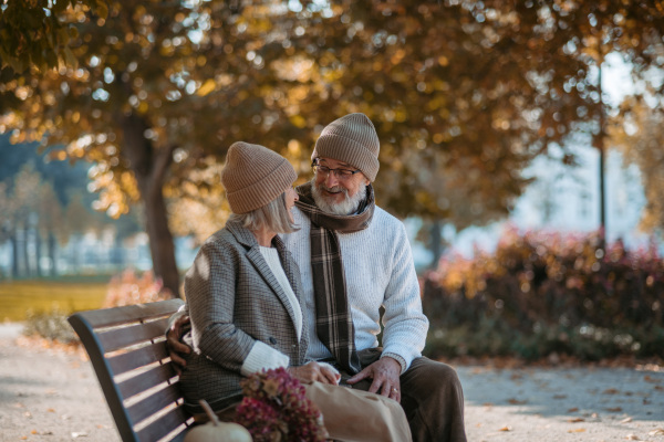 Beautiful senior couple sitting on bench in autumn park. Elderly husband and wife hugging, enjoying fall atmosphere.