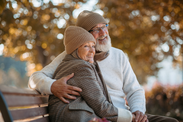 Beautiful senior couple sitting on bench in autumn park. Elderly husband and wife hugging, enjoying fall atmosphere.