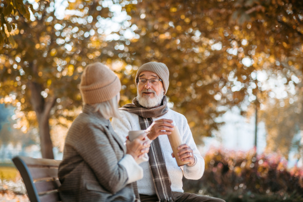 Beautiful senior couple sitting on bench in autumn park. Elderly husband and wife enjoying cup of hot coffee from thermos.