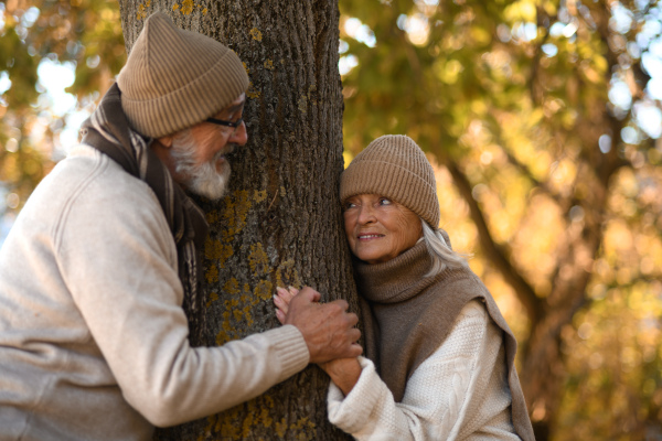 Beautiful senior couple during walk in autumn park. Elderly husband and wife standing by tree trunk, looking at each other lovingly.