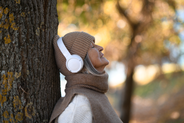 Portrait of a beautiful senior woman with headphones in autumn park, listening music with closed eyes.