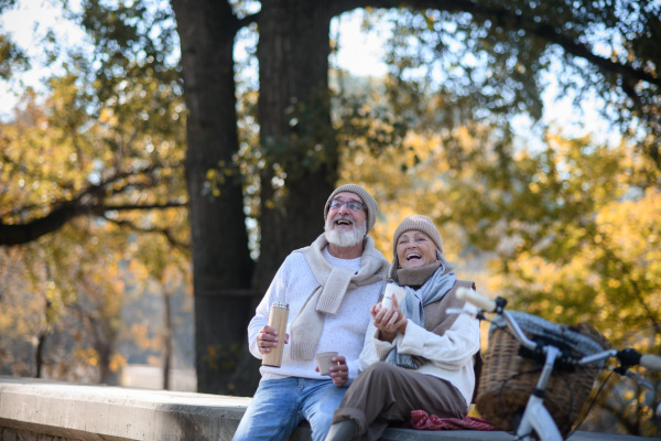 Beautiful senior couple sitting on bench in autumn park. Elderly husband and wife enjoying cup of hot coffee from thermos.