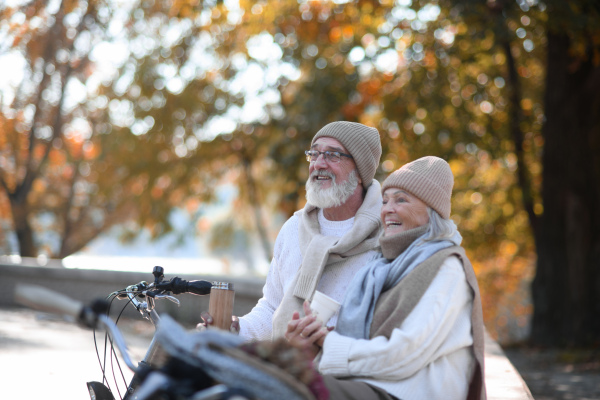 Beautiful senior couple sitting on bench in autumn park. Elderly husband and wife enjoying cup of hot coffee from thermos.