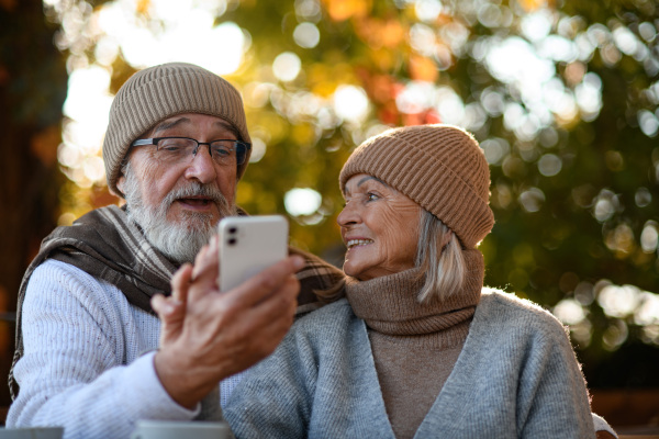 Elderly married couple sitting on coffee shop terrace and taking selfie. Seniors enjoying cup of coffee in the autumn atmosphere.