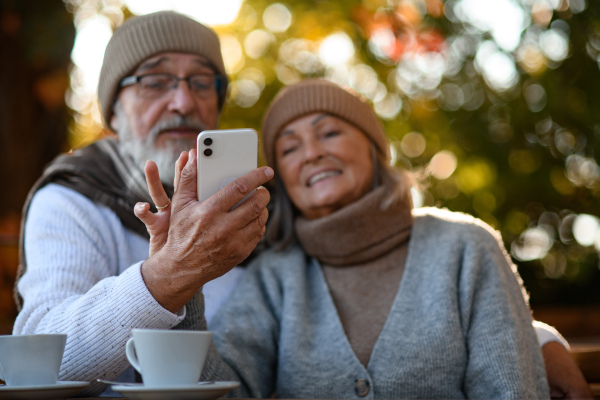 Elderly married couple sitting on coffee shop terrace and taking selfie. Seniors enjoying cup of coffee in the autumn atmosphere.