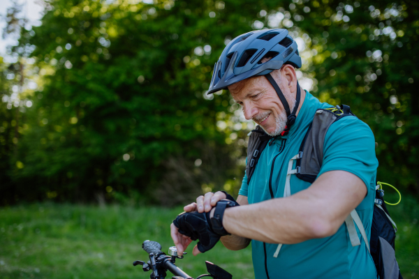 An active senior man riding bicycle at summer park, looking at smartwatch and checking his performance, healthy lifestyle concept.