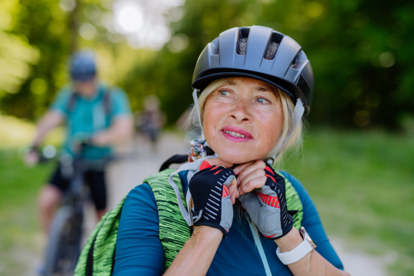 An active senior couple riding bicycles at summer park, woman is putting on helmet, healthy lifestyle concept.